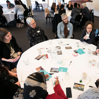 Group of people seated around a round table with notes and coffee cups, engaged in discussion at a workshop.